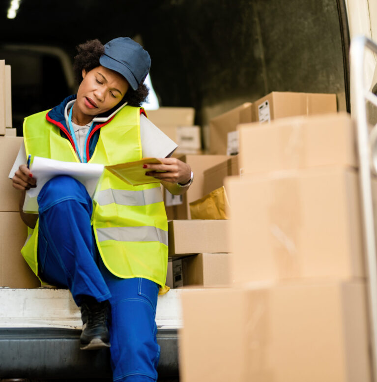 African American delivery woman talking on the phone while going through delivery list in a van.