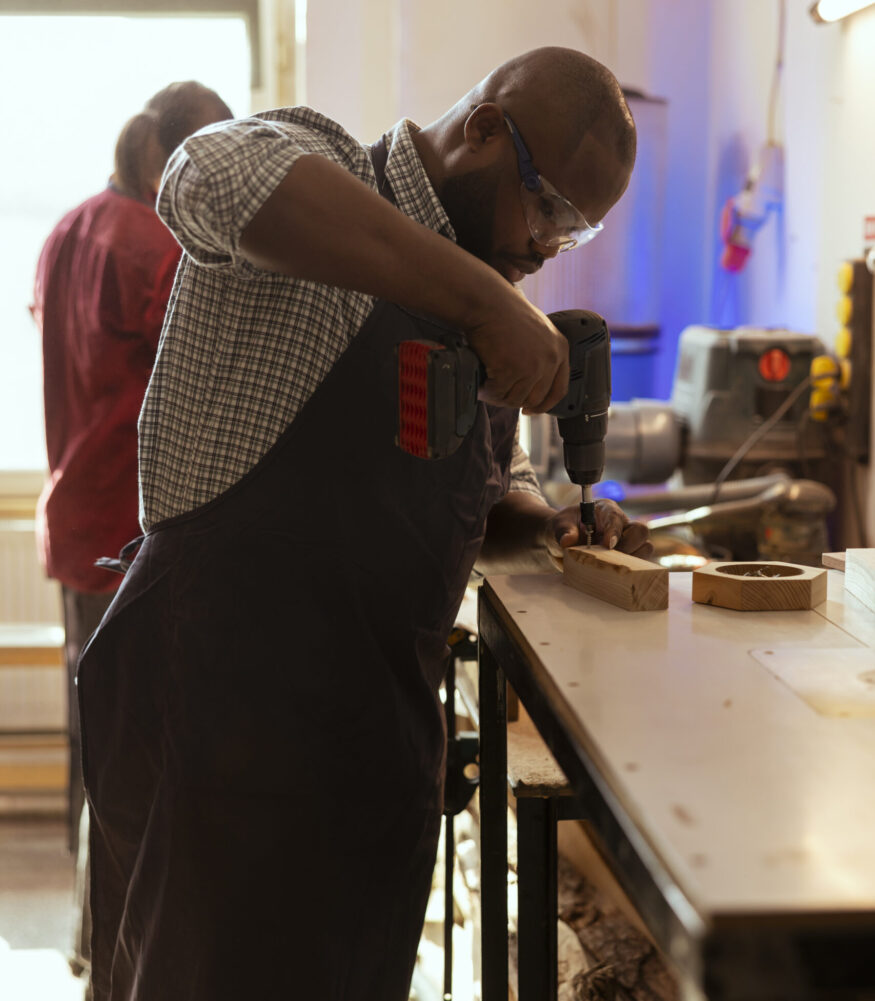 African american carpenter drills hole into timber block, doing furniture assembling in studio. BIPOC woodworking specialist inserting nail into piece of wood using electric power drill tool